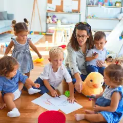 Eine Tagesmutter wird, umgeben von spielenden Kindern. Sie malt mit ihnen auf dem Boden, während ein Stoffentier zusieht.