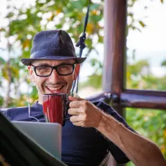 Mann mit Hut und Brille genießt Kaffee im Home Office English, mit Laptop und Kopfhörern.