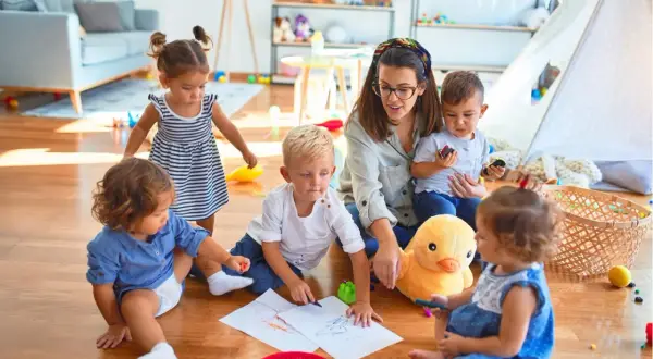 Eine Tagesmutter wird, umgeben von spielenden Kindern. Sie malt mit ihnen auf dem Boden, während ein Stoffentier zusieht.