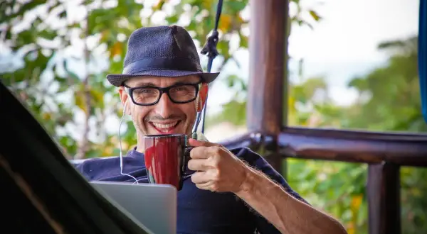 Mann mit Hut und Brille genießt Kaffee im Home Office English, mit Laptop und Kopfhörern.