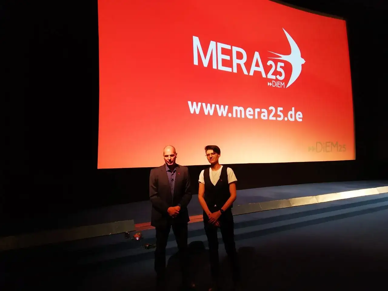 Zwei M&auml;nner stehen vor einer roten Leinwand mit dem Logo von MERA25. Die Leinwand zeigt die Website www.mera25.de. Die Kritik an Diem25 wird hier vielleicht thematisiert.